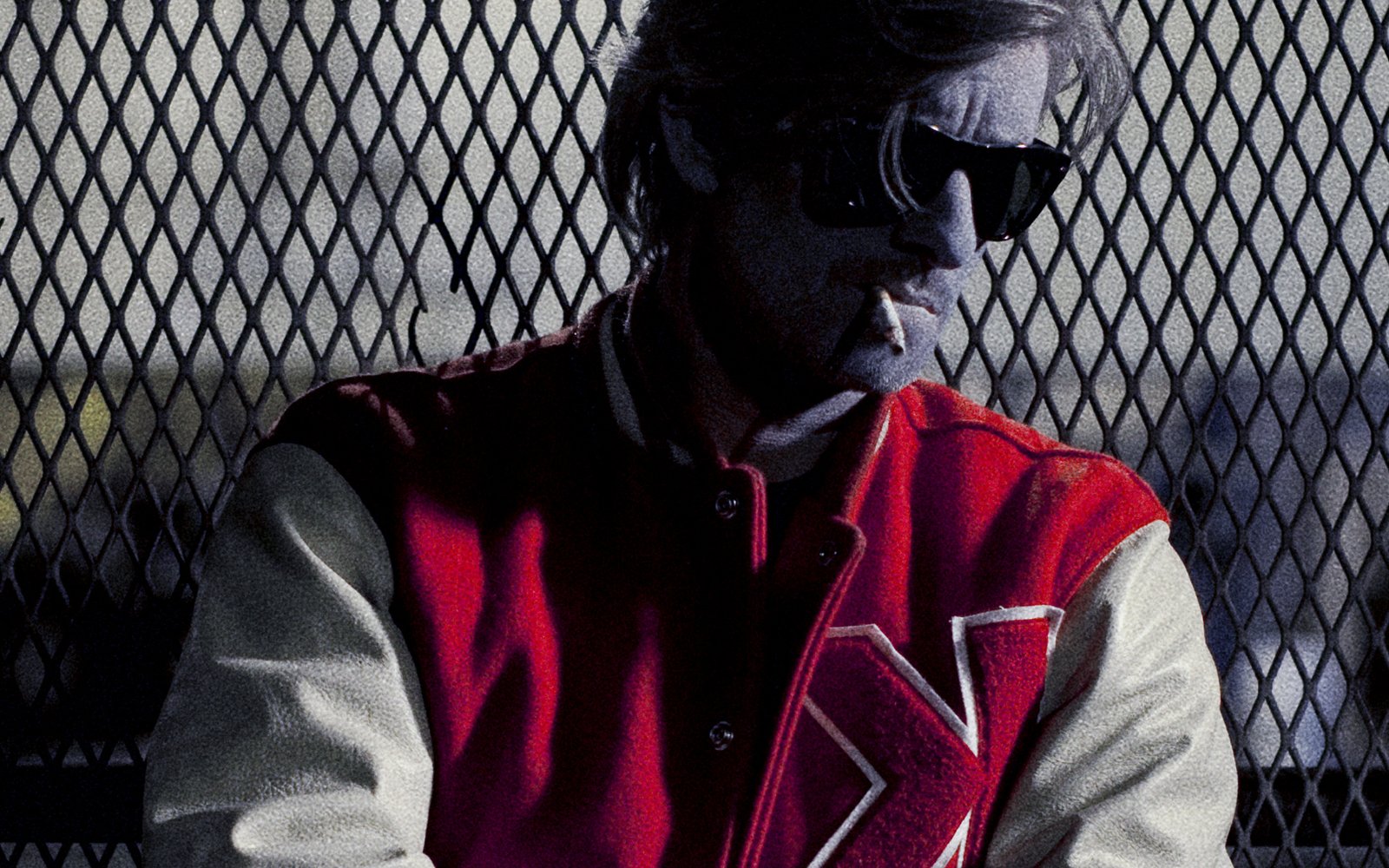 Kavinsky, lunettes noires et blouson rouge au K, adossé à un grillage de nuit